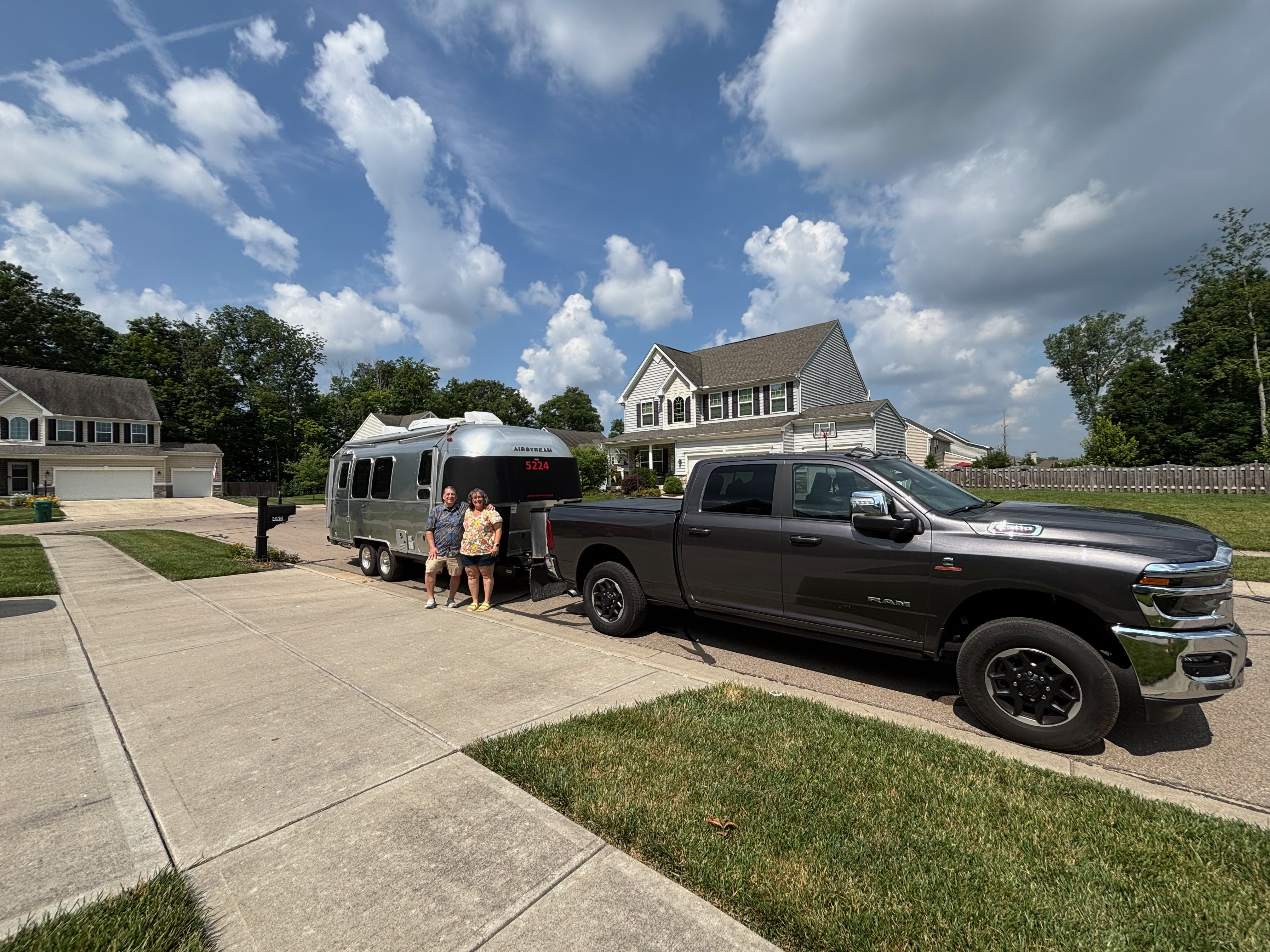 Epsilon, a silver Airstream Globetrotter, hitched behind Smaug, a dark grey Ram 2500, parked across a suburban driveway under a blue sky with tall summer clouds