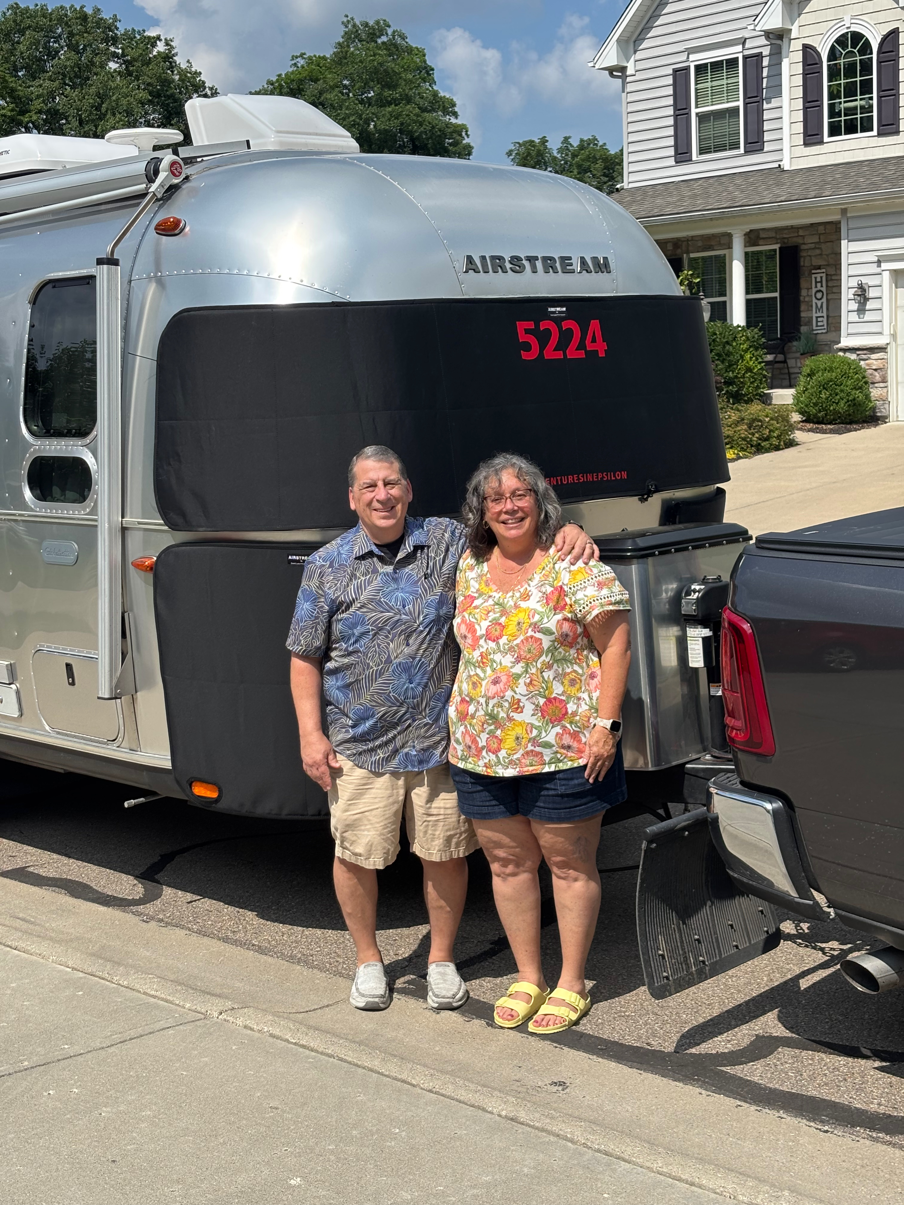 Steve and Colleen standing together in front of Epsilon's nose, the Airstream's stone guard marked 5224, with the truck's tailgate at right