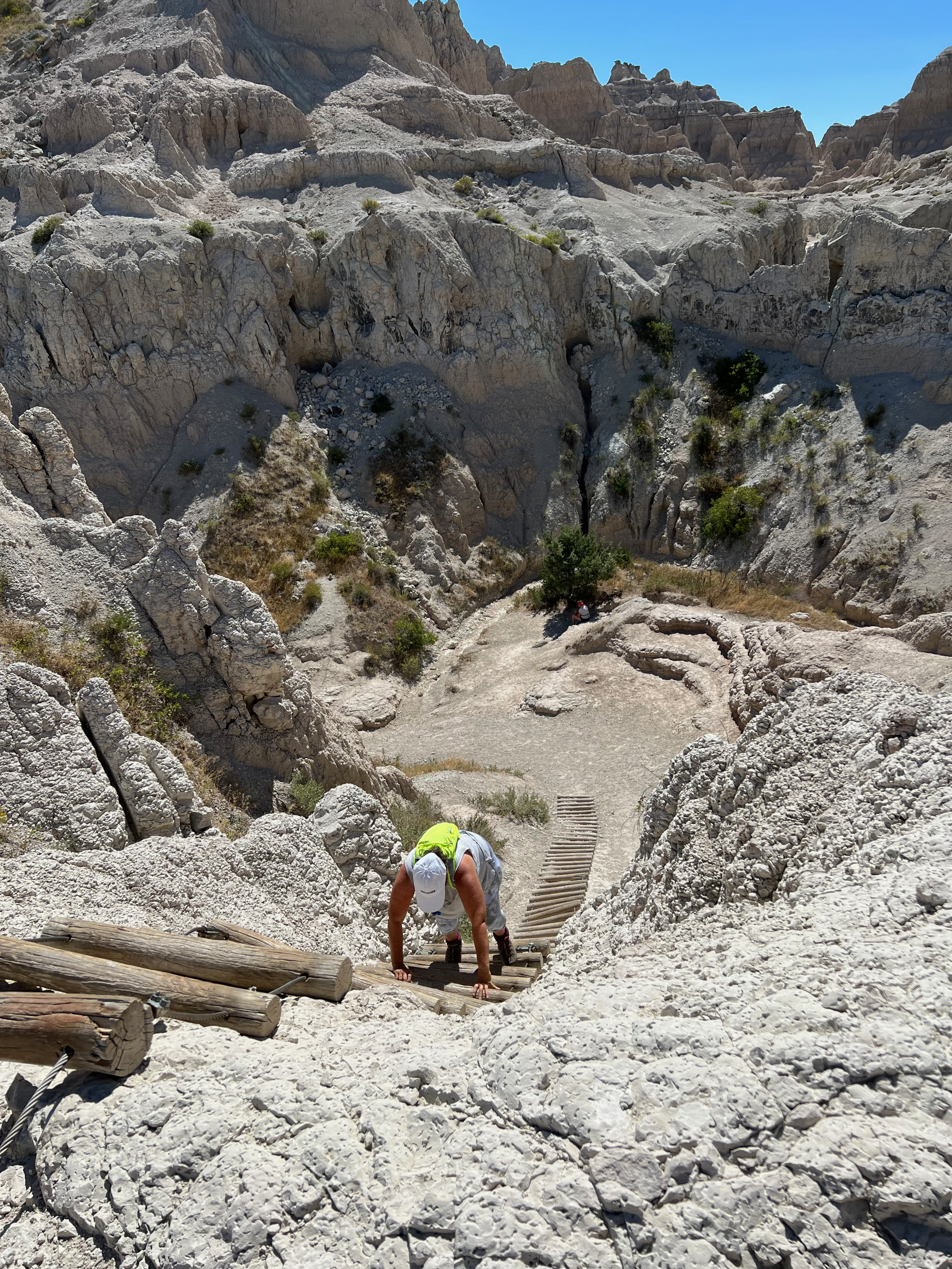 A hiker climbing the wooden log ladder on Notch Trail in Badlands National Park, with eroded white rock formations rising behind
