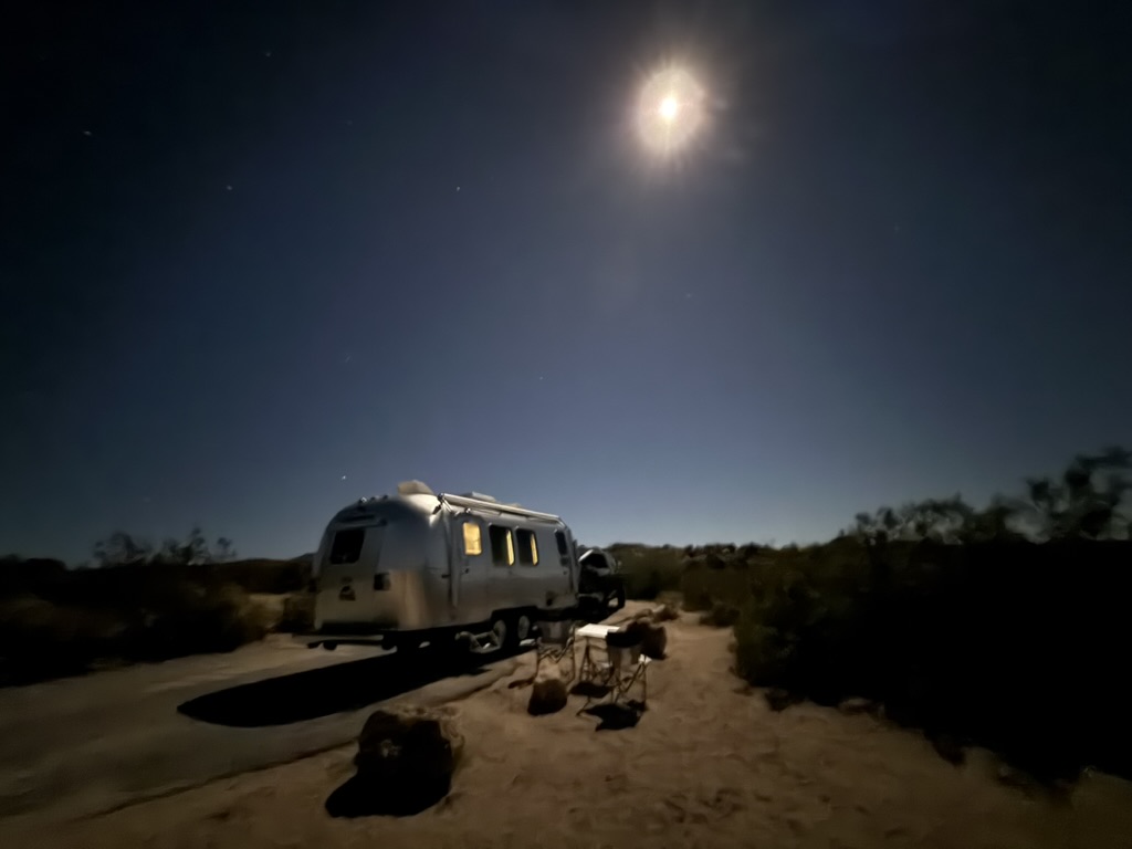 Epsilon, our Airstream, parked in the desert under a bright full moon with camp chairs set up outside