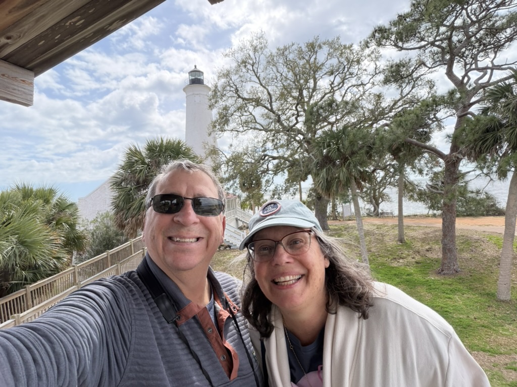 Steve and Colleen smiling in a selfie with the white St. Marks Lighthouse and palms behind them